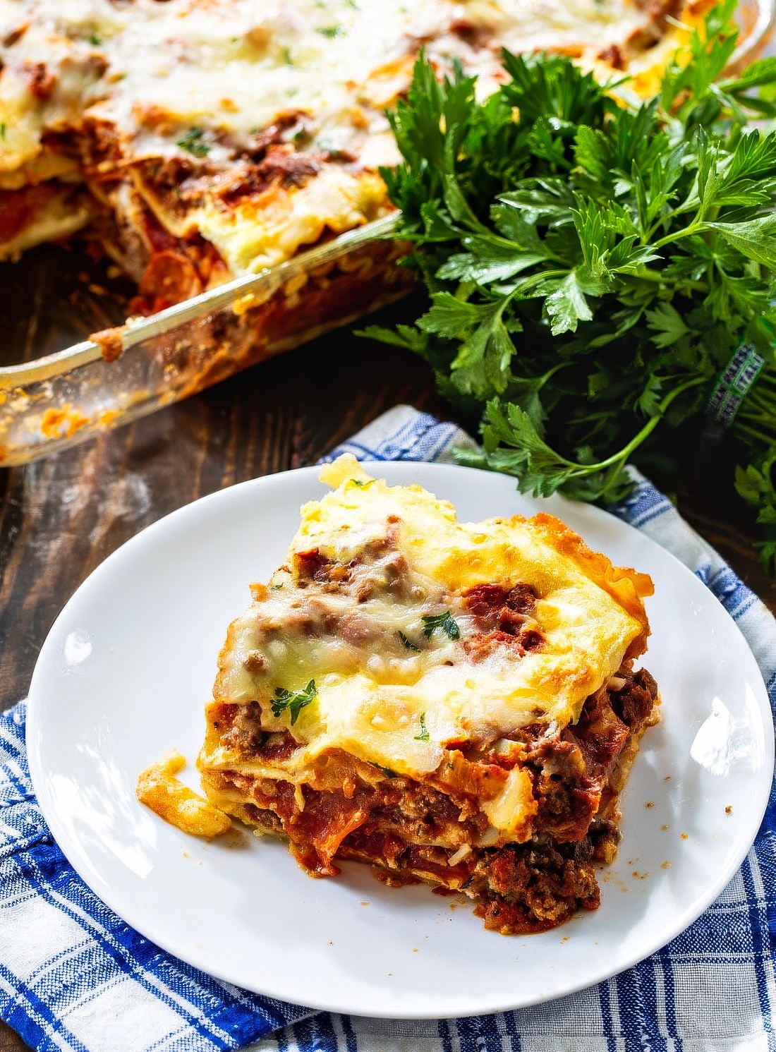 Slice of lasagna on a plate with parsley behind it.