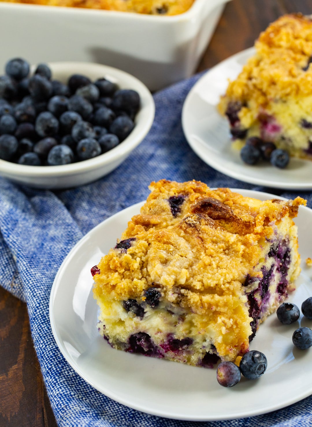 Two Slices of coffee cake on small plates and a bowl of blueberries.