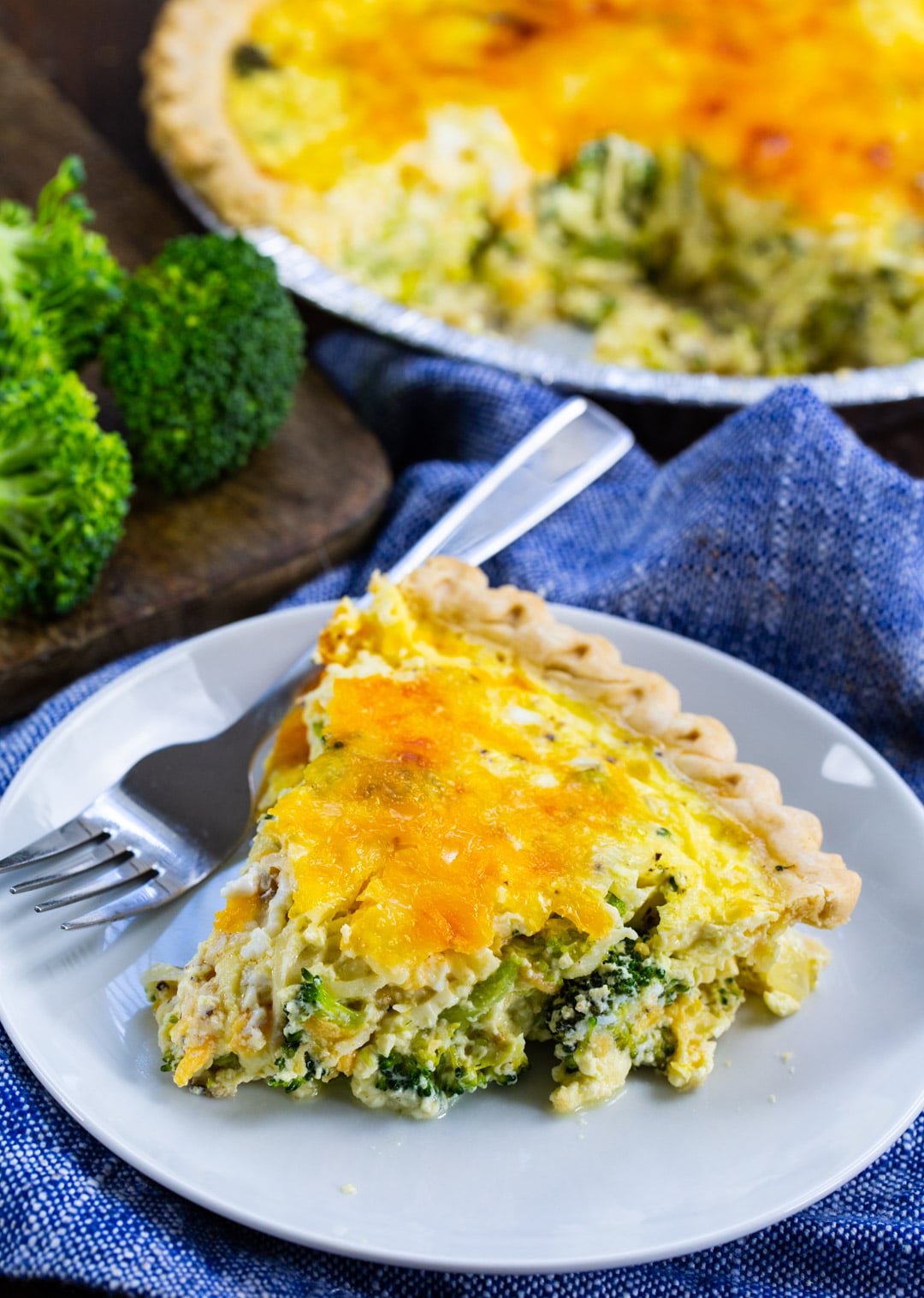 Slice of quiche on a plate with rest of quiche in background.