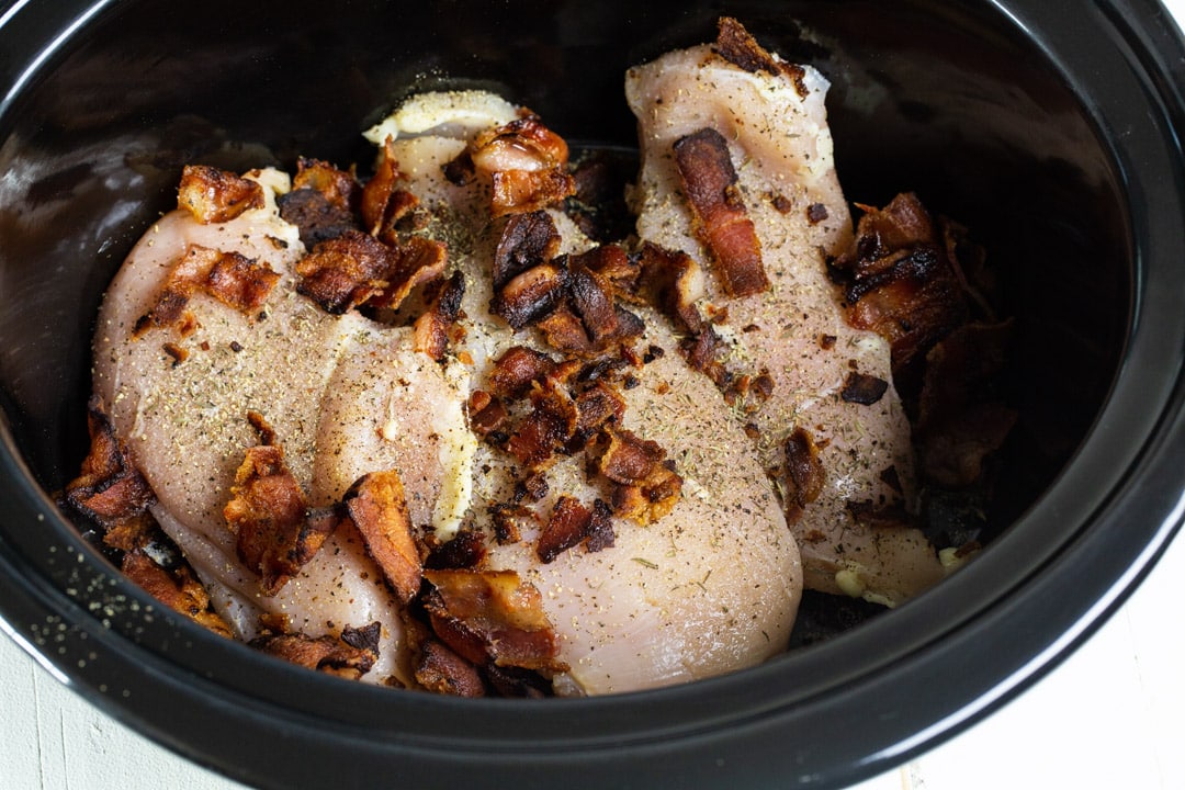 Uncooked chicken topped with bacon in slow cooker.
