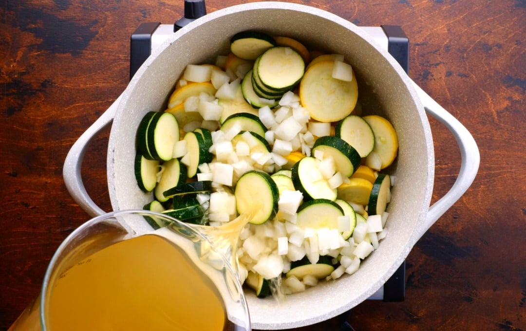 Chicken broth being poured into pot with squash, zucchini, and onion.