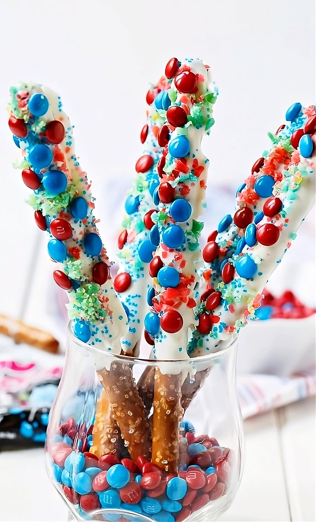 Pretzel rods coated in white chocolate and covered with sprinkles and m&ms.