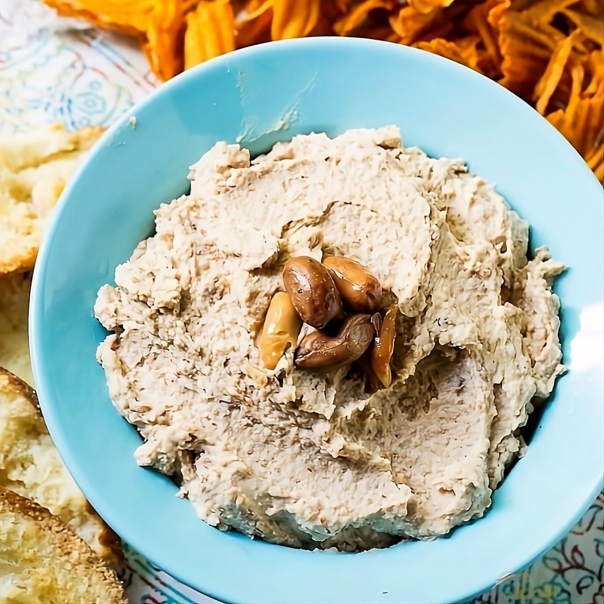 Spicy Boiled Peanut Hummus in blue bowl surrounded by sweet potato chips.