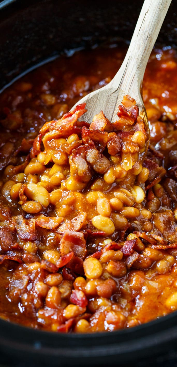 Baked beans in a slow cooker being scooped up with a wooden spoon.