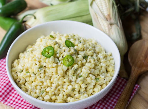 Jalapeno Creamed Corn in white bowl with corn on the cob in background.