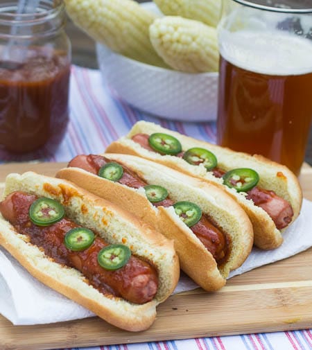 Hot Dogs on a wooden cutting board with beer and corn in background.