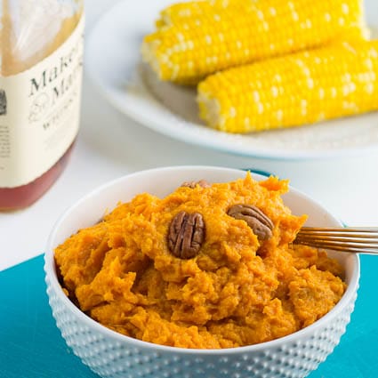 Bourbon Mashed Sweet Potatoes in a white bowl with a bottle of bourbon and corn in the background.
