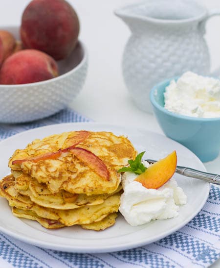 Sweet Peach Pancakes on a plate with fresh whipped cream and a bowl full of peaches in the background.