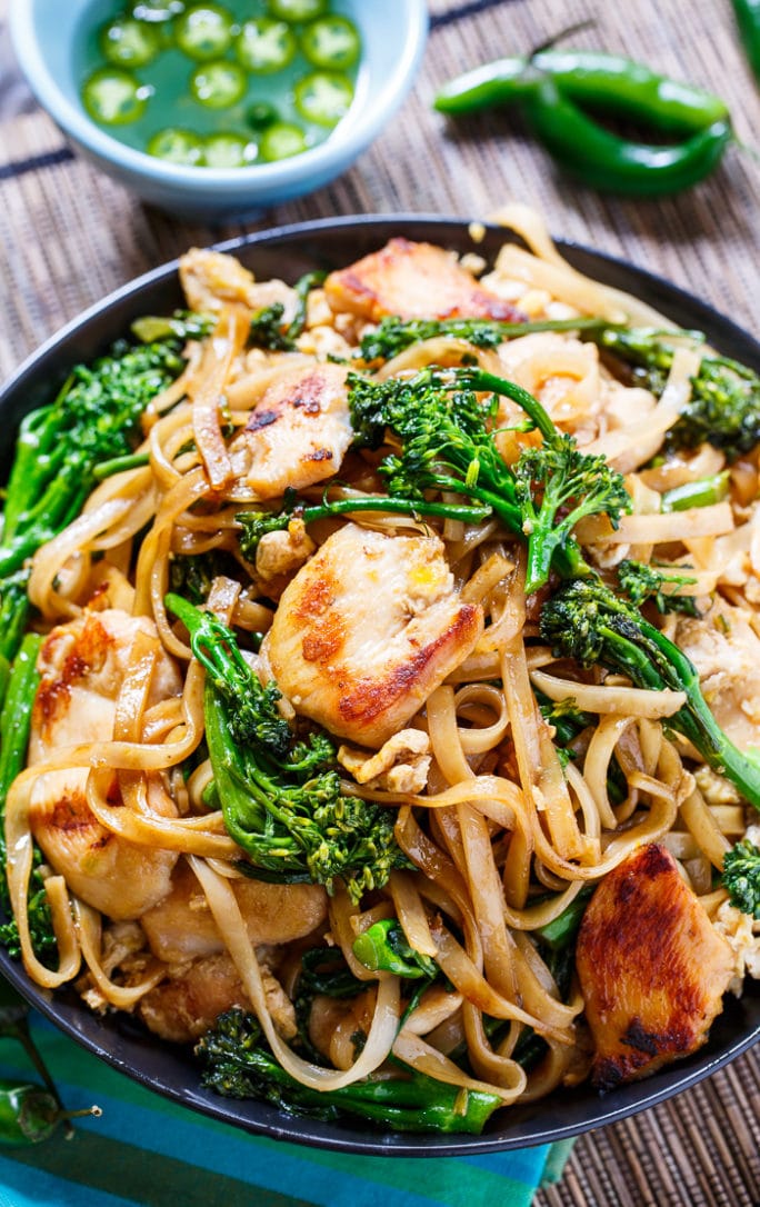 Thai Stir-Fried Noodles with Chicken and Broccolini in a black bowl with jalapenos in background.